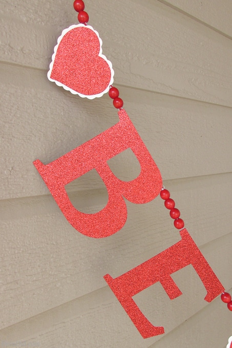 Get ready for Valentine's Day parties and events with this glitter garland inspired by Pottery Barn Kids. This easy Knock Off DIY craft uses glitter cardstock, some twine, and wooden beads to make a charming Valentine's Day banner for under $3.