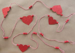 Get ready for Valentine's Day parties and events with this glitter garland inspired by Pottery Barn Kids. This easy Knock Off DIY craft uses glitter cardstock, some twine, and wooden beads to make a charming Valentine's Day banner for under $3.