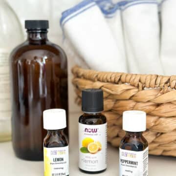 Lavender, lemon, and peppermint essential oil bottles for cleaning on countertop next to cleaning cloths.