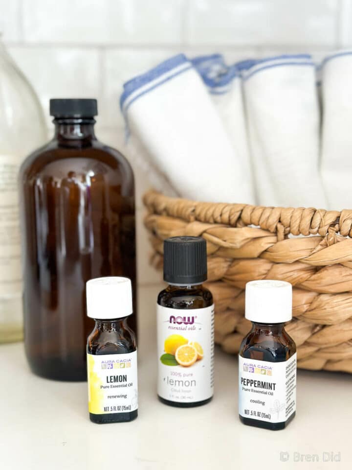 Lavender, lemon, and peppermint essential oil bottles for cleaning on countertop next to cleaning cloths.