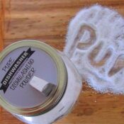 A clear glass mason jar filled with white homemade dishwasher detergent powder on a table and a metal spout with powder poured spelling "pure" featuring a printable label