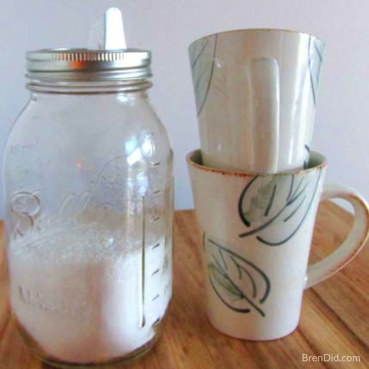 A clear glass mason jar filled with white homemade dishwasher detergent powder, featuring a printable label and a clean mugs resting nearby.